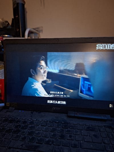 A black Acer laptop with "1080 FHD" branding is depicted indoors, displaying video content. The laptop's keyboard and surrounding chassis are visibly covered in a layer of dust. A dark, slender, rectangular object rests on the keyboard to the right of the touchpad area. The screen shows a scene featuring a male individual with dark hair and glasses, wearing a light-colored collared shirt, looking towards the right side of the frame. Chinese subtitles are present, with an English translation reading "Even if the stone can be lit". Additional Chinese text is visible above and below this line, and in the upper right corner of the screen. The background of the video shows indistinct elements, including a luminous blue screen to the right of the male figure. Above the laptop, a plain light-colored wall is visible, with a dark, vertical rectangular object and other indeterminate items partially visible to the upper left. This scene is located in Überlingen, Germany.