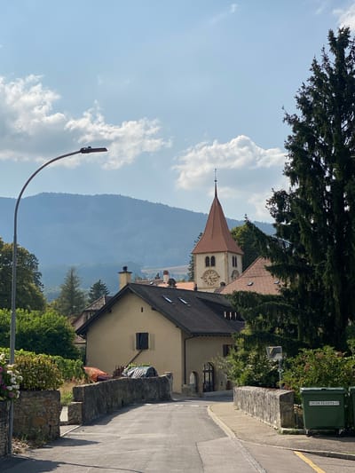 The image shows a street scene in the town of Milvignes, Switzerland, on a sunny day with scattered clouds. A paved road curves to the right, bordered by stone walls. On the left, a lamppost with an LED light fixture arches over the road. Beyond the wall on the left, there are blooming flowers and foliage. On the right, the road continues past a stone wall and two large green recycling bins, one of which has white lettering that reads "CROIX 11-13-15-17".
In the middle ground, several buildings are visible. A light-colored house with a dark roof and a single dark window is prominent. Behind it, a church tower with a red conical roof, a clock face, and a weather vane rises above other buildings. Large evergreen trees are on the right side of the frame, partially obscuring the church tower. In the background, rolling hills covered in trees recede into the distance under a hazy sky. The scene appears to be a quiet, residential area.
