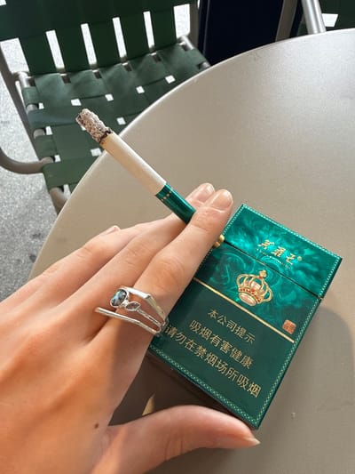 A person is holding a lit cigarette in their left hand. The cigarette is partially burned, with ash visible on the tip. Resting on the person's fingers is a pack of green and gold cigarettes with Chinese writing. The person is wearing a ring with a blue stone on their finger. The scene appears to be outdoors, on a patio or balcony, with a chair with green straps visible in the background. The exact location in the city of Hongkou District, China, cannot be determined from the provided image.