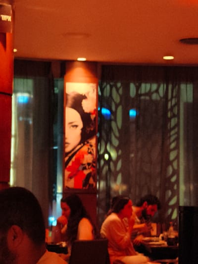 The image depicts an indoor scene, likely a restaurant or bar, with an orange and warm lighting scheme. In the foreground, the back of a person's head and shoulder are visible, suggesting they are seated at a table. Further back, a woman with dark hair is seated, looking towards her right, with her hand near her mouth. To her right, a man with a beard is also seated, appearing to be engaged with something on the table in front of him. The setting features a decorative screen or partition with an abstract pattern behind the seated individuals.
On the left side of the image, a tall, cylindrical structure is partially visible, with a sign at the top displaying Hebrew text. The text reads "אזור" which translates to "Zone" or "Area." To the right of this structure, a framed artwork is displayed, featuring the upper half of a woman's face with dark hair and dramatic makeup, alongside floral elements in shades of red and black. The overall mood is relaxed and intimate, typical of an evening setting. No specific weather conditions are discernible. The presence of the Hebrew text suggests the location is in Israel, and the prompt specifies Be'er Sheva.