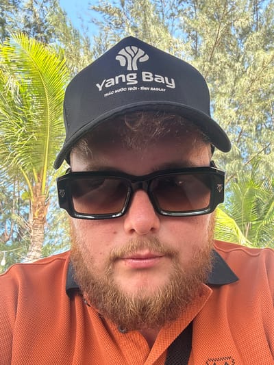 A close-up, over-the-shoulder selfie shows a fair-skinned person with a beard and ginger hair wearing a black baseball cap and black sunglasses. The cap has a white logo and text that reads "Yang Bay Thác Nước Trời - Tỉnh Raglay". The person is wearing an orange collared shirt. The background consists of palm trees and foliage, consistent with a tropical environment. The image was taken in Ninh Chu Ward, Vietnam.