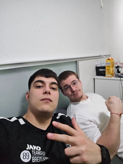The image depicts two males inside a residential room in Krems an der Donau, Austria. In the foreground, a male with dark, short hair and a neutral expression looks directly at the camera. He wears a black t-shirt with white shoulder stripes and the text "JAKO TEAMSHOP 89 BREITLESS." His right hand is raised, fingers forming a 'V' gesture, and he wears a dark wristband on his left wrist. Positioned behind him to the right, a second male with light brown hair, a beard, and eyeglasses also looks towards the camera with a slight smirk. He wears a plain white t-shirt and a red beaded bracelet on his left wrist, with his left arm bent and hand forming a fist. The background consists of a plain white, textured wall and a white cabinet. On the cabinet, a yellow cardboard carton, a black container with white material, a black electronic device, and a white power cord are visible. A grey horizontal trim piece runs along the wall.