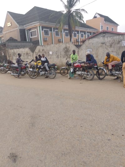 The image captures a daytime scene in Ijaiye, Nigeria, showing a group of men gathered around several motorcycles on a paved road next to a low concrete wall. At least eight men are visible: some are standing beside their motorcycles, while others are seated on them.
On the left, a man in a plaid shirt stands near a red motorcycle, with another man in a white and black jacket seated on a yellow motorcycle nearby. Towards the center, a man in a light green top stands by a motorcycle, and another in a blue hoodie is hunched over a red motorcycle. Further right, a man in a yellow top and light-colored pants sits on a motorcycle. The men appear to be resting, waiting, or socializing.
In the background, a large, multi-story building with a distinctive dark roof and light-colored walls with green trim is visible. Several air conditioning units are mounted on its exterior. A tall palm tree stands to the right of the building. The sky is bright but appears overcast or hazy, suggesting either early morning or late afternoon. The overall setting is consistent with an urban or semi-urban area. No visible text is discernible in the image.