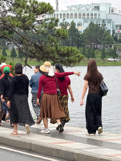 In Xuan Huong Lake, Da Lat, Vietnam, a group of six individuals, predominantly women, are observed walking along a paved pathway adjacent to the water. The scene is dominated by a large, light-colored, multi-story building with arched windows in the background, characteristic of the architecture found in Da Lat. Pine trees line the path and the grounds of the building, adding to the naturalistic setting.
The individuals are dressed in casual, layered attire, suitable for a temperate climate. Several are wearing hats, including a straw hat with a large bow and a green baseball cap. Their clothing consists of skirts, dresses, and trousers in various colors and patterns, such as polka dots and floral prints. One woman, facing away from the camera, gestures towards the right with her arm outstretched. Another woman, also facing away, carries a black leather handbag. The ground is a tiled walkway with a textured surface, bordered by a concrete curb. A body of water, presumably Xuan Huong Lake, stretches along the right side of the frame, reflecting the overcast sky. A white vehicle is visible on a road in the distance, beyond the lake's edge.