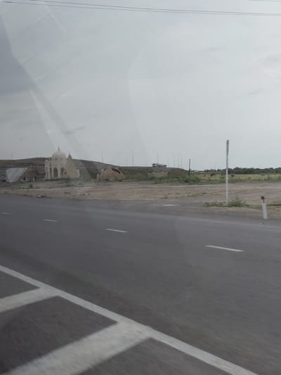 The image depicts a roadside scene with a prominent, light-colored dome-shaped structure in the distance, possibly a mausoleum or a shrine, located in an arid or semi-arid landscape. Adjacent to it are several ruined or incomplete mud-brick structures. A paved road with white lane markings runs diagonally across the foreground, suggesting a current or recent movement of the camera, likely from a vehicle. The sky is overcast, casting a diffused light over the scene. In the background, there are some distant buildings or infrastructure, including streetlights, and a truck can be seen on a road further away. The overall environment appears somewhat desolate with sparse vegetation. The location is identified as Нарын ауылдық округі, Kazakhstan.