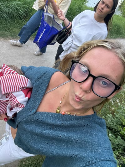This media file captures a casual outdoor scene featuring two young women, likely in Marquette-lez-Lille, France. The image is taken from a selfie perspective by the woman in the foreground, who has blonde hair and wears black-framed glasses, a gold nose ring, a blue knitted cardigan, and white pants. She makes a playful pouting face for the camera and accessorizes with multiple gold necklaces, including charms like a shell and a star. She is holding a red and white striped item, possibly a towel or blanket, and her nails are painted red.
Behind her, another woman with dark long hair walks along a paved path. She wears a white t-shirt, grey sweatpants, blue jeans, and white sneakers, and carries a black shoulder bag along with a blue tote bag. The blue tote bag has "grandfrais.com" printed on it, which is a French supermarket chain.
The setting appears to be a paved pathway running alongside a body of water, possibly a canal or river, with lush green grass and dense foliage on the banks. The lighting suggests it is daytime, perhaps an overcast day or late afternoon. The overall atmosphere is relaxed and informal, capturing a moment during an outdoor stroll.