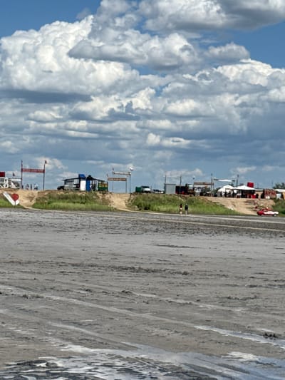 The image captures a scene at the Solonchak Terek-Tereze in Bursol', Russia, characterized by a vast expanse of dried mud flats in the foreground. In the mid-ground, a dusty embankment hosts a collection of structures, including tents, a small building with a blue roof, and various stands or stalls. Several vehicles, including a red classic car, are present. People are visible walking on the embankment and on the mud flats. Flags, some bearing Russian colors, are mounted on poles above some of the structures, indicating a public gathering or event. The background is dominated by a dramatic sky with large, puffy cumulus clouds against a blue sky. The overall environment appears dry and open, consistent with a salt flat or similar geological formation.