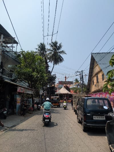 This image depicts a bustling street scene in Kochi, Kerala, India, under clear daylight. The viewpoint is from a slightly elevated position, looking down the asphalt road.
In the foreground, a person wearing a white helmet and a light blue checkered shirt rides a dark-colored scooter (license plate KL-63-L-XXXX, partially visible) away from the viewer, centered on the road. To the right, a dark blue Maruti Suzuki Omni van (license plate KL 43 D 2049) is parked parallel to the curb, with another scooter partially visible in front of it.
The left side of the street features a narrow sidewalk fronting a row of commercial establishments or residences. Multiple scooters and motorcycles are parked along this sidewalk. Figures are vaguely discernible near the storefronts. Overhead, numerous electrical and communication cables crisscross the street and connect to utility poles and buildings.
In the midground, an auto-rickshaw (yellow and black) and another light-colored scooter are visible further down the road. A light-colored utility truck is parked on the right side of the street, alongside the blue van.
The background reveals a large decorative archway or gateway, possibly a temple entrance, adorned with red and yellow elements, which serves as a focal point at the end of the visible street. In front of this gateway, a prominent light-colored, conical tent structure is erected. Buildings frame the street: on the left, a multi-story building with a flat roof and balconies, characterized by exposed utility wiring; on the right, an older, two-story structure with a corrugated metal roof and a faded light brown facade, its lower wall covered with posters. Several palm trees and other green foliage are visible against a clear, light blue sky. Decorative string lights are strung across the street in the distance.