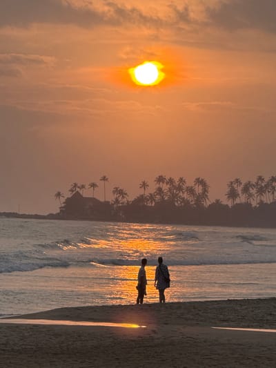 The image depicts a sunset or sunrise scene on a beach in Dikwella, Sri Lanka. The sky is dominated by an intensely bright, circular white and yellow sun, centered high in the frame, casting an orange and amber glow across scattered clouds and the atmosphere. Below, two silhouetted individuals stand in shallow water or on the wet sand near the ocean's edge, facing away from the camera and towards the illuminated sea.
The individual on the left appears to be male, wearing a short-sleeved top and shorts, with a slight turn of the head towards the right. The individual on the right appears to be female, dressed in a patterned garment, possibly a dress, and carrying a bag over the left shoulder. Both figures are stationary, observing the ocean.
The sea exhibits gentle waves, with the water reflecting the sun's bright orange light across its surface in a shimmering path towards the foreground. The immediate foreground consists of wet sand on the beach, also reflecting the sun's glow. In the mid-ground, across the water, a distant landmass or island is visible, densely covered with numerous palm trees that are silhouetted against the lighter sky. A building, possibly a structure with a peaked roof, is discernible amidst the palm trees on the left side of this distant landmass. A rocky breakwater or natural rock formation extends into the water from the left side of the distant landmass.