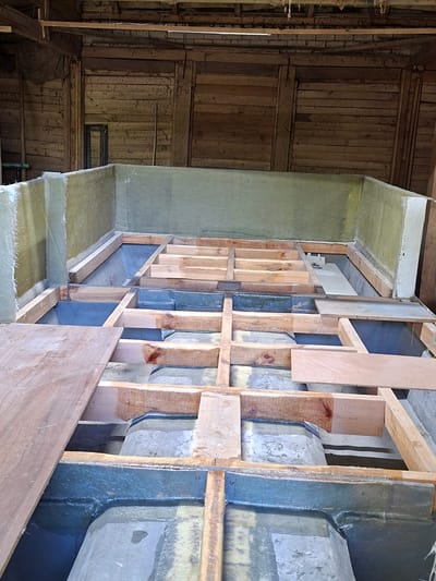 The image depicts the interior of a wooden structure, possibly a barn or workshop, undergoing construction or renovation. The foreground is dominated by a partially constructed fiberglass hull or tank, with a wooden support structure built around and over it. The hull is a light green color with a rough texture, and the wooden supports consist of various planks and beams arranged in a grid pattern. Some sections of the hull are exposed, revealing a dark blue or gray interior. The background shows rough-hewn wooden walls and ceiling, with exposed beams and planks. The overall impression is one of a construction site, with unfinished elements and raw materials. The location is stated to be in the city of Žemaitkiemis, Lithuania. There are no people visible in the image.
