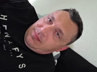 A portrait of an adult male, located in Shotton, United Kingdom, captured from an elevated, oblique angle. The individual has fair skin, dark, short hair with visible graying at the temples, and a receding hairline. His hazel-toned eyes are directed at the viewer. Horizontal creases are present on his forehead. A distinct, raised brown mole is visible on his left cheek. Short facial stubble covers his chin and jawline. His lips are closed, forming a subtle, relaxed expression. He is wearing a black garment, likely a t-shirt or sweatshirt, featuring white text 'HENLEYS' across the chest. The background consists of a light-colored, textured wall to the upper right and behind his head. A light green textile is visible behind his upper left shoulder, and a gray and white striped textile is present beneath his chin.