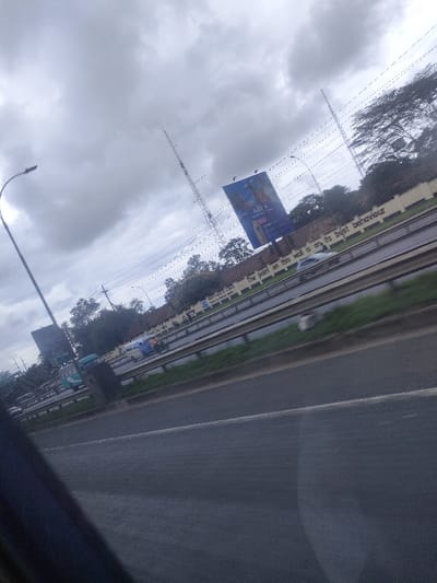 The image displays an overcast daytime scene of a multi-lane highway in Juja ward, Kenya. The perspective is angled diagonally, suggesting observation from a moving vehicle, with a dark, indeterminate object partially obscuring the lower left foreground.

The highway features at least four visible asphalt lanes, separated by a grassy median strip and metal guardrails. Multiple vehicles, including cars and what appear to be buses, are in motion on the roadway, some exhibiting motion blur. A light-colored sedan is discernible on an inner lane.

Adjacent to the highway, a light-colored retaining wall displays the painted text: "THE POINT ON THIS WALL IS ON ITS BEST BEHAVIOUR." Behind this wall, a dense line of trees and foliage is visible.

Two tall communication or utility towers punctuate the mid-ground and background, rising against a uniformly gray, heavily overcast sky. Numerous overhead power lines or cables traverse the upper portion of the frame. Two large billboards are present: one prominently features an advertisement including the text "48 Laws" and images of individuals and a product container; another less distinct billboard is situated further left. A streetlamp with a curved arm stands on the far left. No discernible individuals are present outside of vehicles.