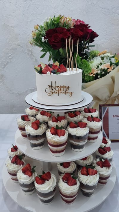 A tiered dessert stand displays a white birthday cake and individual layered trifles topped with strawberries and cream. The cake, adorned with gold candles and fruit, is situated on the top tier, while the trifles are arranged on two lower tiers. The backdrop features a bouquet of red roses and other flowers against a textured white wall. A framed document with text in Cyrillic script is visible to the right. The scene appears to be set indoors on a table.