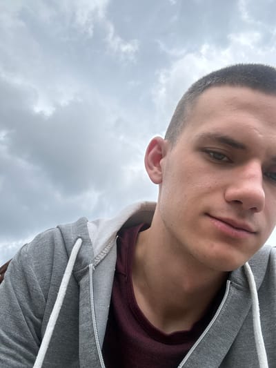 A close-up, low-angle, selfie-style shot features a young man with short, dark hair and fair skin. He is wearing a gray hooded sweatshirt over a maroon t-shirt. His head is slightly tilted, and his gaze is directed downwards, with his right eye visible. The background consists of a cloudy sky. The image appears to have been taken outdoors in Momchilovtsi, Bulgaria.
