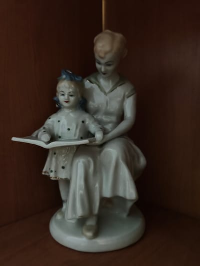 A porcelain figurine depicts a woman and a child sitting together, with the woman's arm around the child. The child is holding an open book. Both figures are dressed in white. The figurine is displayed on a wooden surface, likely a shelf, against a dark wooden background. The lighting is dim, casting shadows. There is no information available about the location of this image in Bucha, Ukraine.