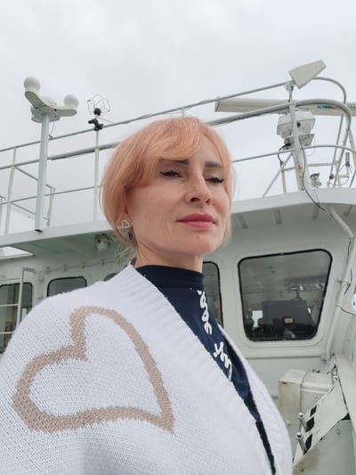 A woman with peachy-blonde hair and makeup poses for a selfie on the deck of a white boat. She is wearing a white cardigan with a large tan heart design on the front and a dark blue shirt underneath. Her left ear is adorned with a small, heart-shaped earring. In the background, the superstructure of the boat is visible, along with various nautical equipment including antennas and a radar dome. The sky is overcast and a building, likely part of the boat's interior, is visible through a window. The boat appears to be docked or stationary, as there is no indication of movement. The location is the city of Vytegra, Russia, likely along the Volga-Baltic Waterway.
