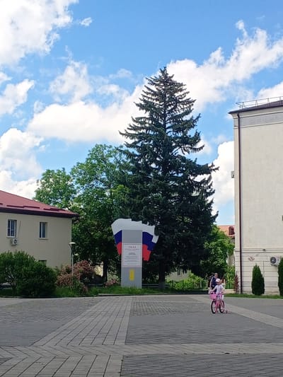 A wide shot captures a square in Yessentukskaya, Russia, on a partly cloudy day. In the foreground, a young girl rides a pink bicycle with training wheels on a paved surface. Behind her, a woman walks. A large evergreen tree dominates the midground, with a monument featuring the Russian flag and text standing to its left. A light-colored building with a red roof is visible on the left, and a taller, light-colored building stands on the right. Green trees and bushes dot the landscape.