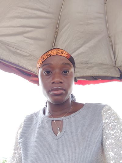 A young Black woman is centered in the frame, wearing an orange patterned headscarf and a gray top with sequined sleeves. She has a silver necklace with a pendant. The woman stands beneath a tent or awning, judging by the fabric visible overhead and to the sides. The background is brightly lit, possibly indicating daylight. The location is potentially in Umuahia, Nigeria, as specified in the prompt. The absence of additional visual information makes it impossible to confirm this location or to elaborate on any specific actions, interactions, or objects within a broader environmental context.
