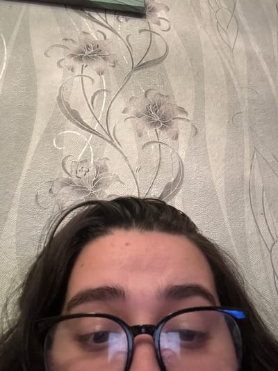 The image shows a close-up view of a person wearing glasses. The person has dark hair and eyebrows. The background consists of a wallpaper with a floral pattern. The location is unknown, and there is no discernible connection to Dobrovnitsa, Bulgaria.