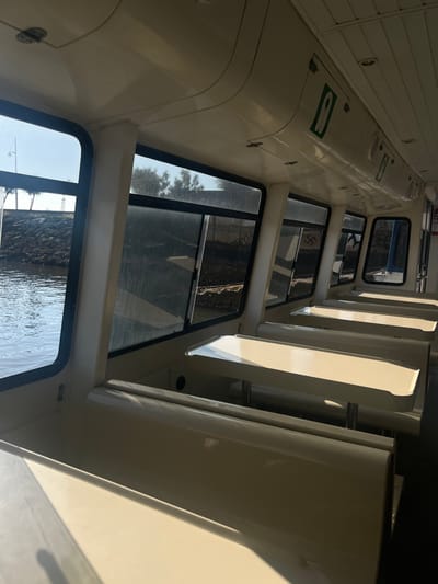 The image depicts the interior of a passenger vessel, likely a ferry, situated in Ribamontán al Mar, Spain. The cabin is characterized by an absence of occupants and features multiple rows of fixed booth-style seating. Each seating unit consists of two opposing benches and a central table, all uniformly light cream in color with rounded edges on the tables. The ceiling and upper wall panels are also light cream, incorporating recessed lighting fixtures and a green emergency exit pictogram. Large, rectangular windows line both sides of the passenger compartment. Through the windows on the port side, calm water is visible adjacent to a dark, rocky embankment, with a solitary light pole and sparse trees in the background under a bright sky. Through the windows on the starboard side, a landmass is apparent, featuring a concrete wall extensively covered in dark graffiti, notably including a visible swastika symbol, with some dark vegetation beyond. The vessel appears to be docked or operating close to a shoreline.