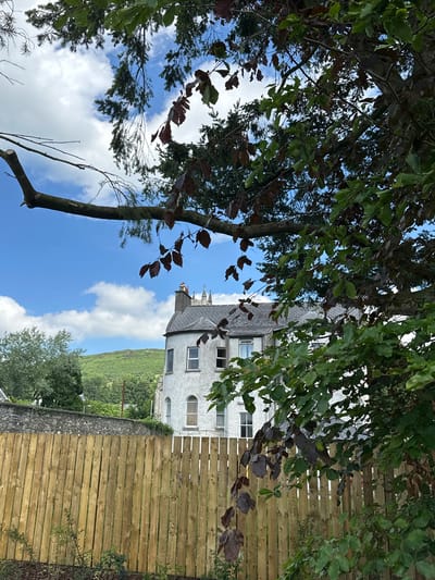 The image depicts an outdoor scene, possibly in Baltinglass, Ireland, with a light gray building featuring multiple windows visible through the branches of leafy trees. The building has a unique architectural style with arched windows and what appears to be a slate roof. In the foreground, a wooden fence runs horizontally across the frame, constructed from light-colored planks. Behind the fence and building, a lush green hillside rises, suggesting a rural or semi-rural setting. The sky is a vibrant blue, dotted with fluffy white clouds, indicating fair weather and possibly daytime. The presence of trees with green and some reddish-brown leaves suggests it could be late summer or early autumn. The perspective is from below, looking up through the foliage towards the building and sky. There are no people or specific activities clearly depicted, giving the scene a quiet and serene atmosphere. The overall impression is one of a tranquil landscape with a blend of natural elements and man-made structures.