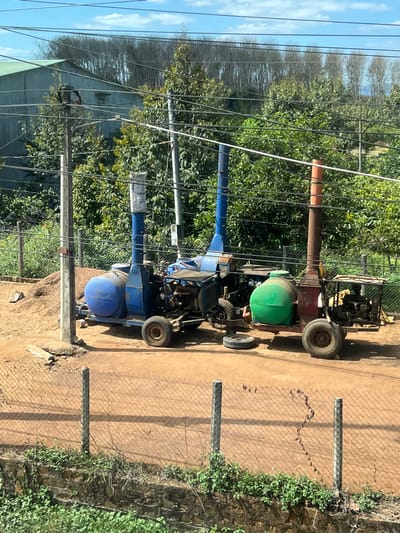 Three farm vehicles sit on a dirt path next to a chain-link fence. Two are blue with cylindrical tanks, while the other is red and green. Trees and utility poles with visible power lines fill the background. A building is partially visible on the left. The path runs parallel to the fence, with green vegetation visible on the other side.