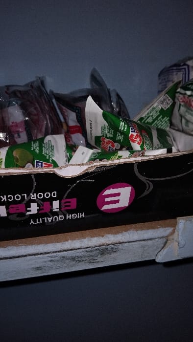 The image shows a close-up of a shelf with several packages of coconut milk sitting on top. The shelf is made of wood and has a white paint finish. The packages are all wrapped in clear plastic and have green and white labeling. The top of the shelf is partially obscured by the packages. The shelf is located in Kecamatan Panjalu, Indonesia (latitude: -7.143, longitude: 108.278). The shelf is labeled "HIGH QUALITY Eifee DOOR LOCK" and features a pink "E" logo. The shelf appears to be part of a home or shop.  There are no people in the image.