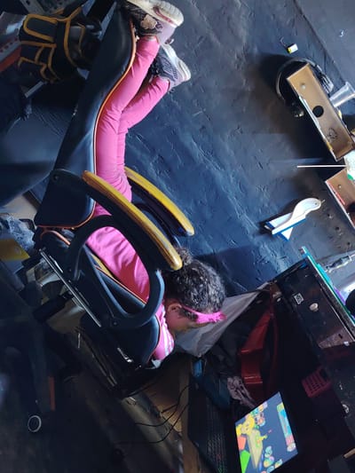 A young girl is reclining in an office chair in a cluttered room. She is wearing pink leggings, a pink and white striped top, and tennis shoes. Her head rests on a white pillow. A computer monitor displaying a game is on a desk near her, along with a keyboard and other items. The room appears disorganized and contains various objects, including an open box, a toy plane, and what looks like disassembled electronics. There is not enough information to determine if the location is Trujillo, Peru.