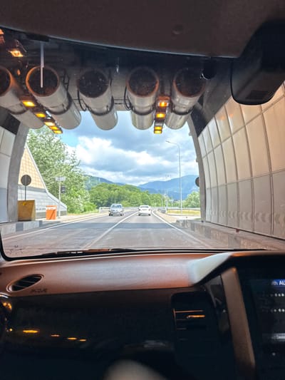 The image captures a view from inside a vehicle, looking out through the windshield. The vehicle is approaching the exit of a tunnel, with daylight and a mountainous landscape visible ahead. Inside the tunnel, directly above the windshield, a series of cylindrical structures with orange lights are mounted on the ceiling. The tunnel walls are white and modular. On the road ahead, two cars are visible, one silver and one white, traveling in the same direction. The right side of the vehicle's dashboard is partially visible in the foreground. This scene is consistent with a road leading out of a tunnel in the Adlerskiy district of Sochi, Russia, known for its mountainous terrain and infrastructure projects.