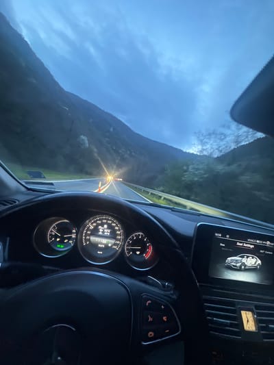 The image shows the interior of a car, specifically the dashboard and steering wheel, while driving on a winding road. The dashboard features a digital display showing "18:58", "VIVACO", "Seat Settings," and a 360° camera view of the car. An analog clock is also visible. The speedometer indicates a speed of 10 km/h. The car's temperature gauges show 103°C and 93°C. The gear selector is in "D". Outside the windshield, a mountain landscape is visible under a cloudy sky. The road ahead is winding with guardrails and orange warning lights visible. There is no indication of the precise location in the city of Mechkul, Bulgaria.