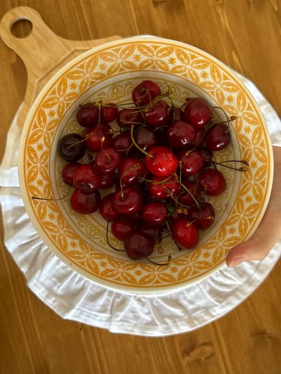 A close-up, top-down shot shows a person's hand holding a yellow-rimmed bowl filled with ripe cherries. The bowl features a decorative pattern of orange floral motifs. The cherries are a deep red color and have stems attached. They are piled in the bowl, with some slightly darker, almost black cherries mixed in. The bowl sits on a white ruffled cloth, which is placed on a wooden surface. To the left of the bowl, a portion of a wooden cutting board with a circular handle is visible. The lighting suggests it is daytime, and the scene appears to be indoors. No visible text is present in the image. The overall impression is one of freshness and natural abundance.