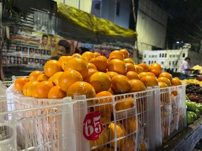 This media file depicts an outdoor market scene at night in Chennai, India. The primary subjects are crates overflowing with bright orange fruits, likely oranges, arranged in plastic white baskets. In the background, a poster with images of several men is visible, suggesting a political or promotional display. Further back, the indistinct shapes of buildings and stalls indicate a bustling marketplace. The lighting is artificial, typical of nighttime street markets, casting a warm glow on the fruits and creating shadows.
The scene appears to be active, with implied commerce taking place, although no direct human activity is clearly discernible in the foreground. The focus is on the vibrant display of produce. One crate in the foreground has a red sticker with the number "65" in white, which may denote a price or quantity. Chinese characters are also visible on one of the baskets. The overall impression is that of a lively, albeit somewhat dimly lit, market environment in an urban setting. The weather appears to be clear, as there are no indications of rain or adverse conditions. The time of day is clearly night, given the artificial lighting and dark background.