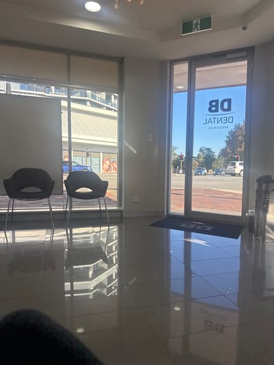The image captures an interior view, likely a waiting area, within a building in Applecross, Australia, as indicated by the “DB Dental” signage reversed on the front window. The scene is observed through two sets of windows, one overlooking a street with vehicular traffic, including cars and a traffic light, and the other revealing urban architecture across the street. Two black chairs are situated in the waiting area, oriented away from the windows towards an unseen area within the building. Reflections are prominent on the tiled floor, mirroring the dental office's branding and exterior view. The composition is a little crooked.