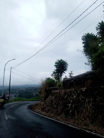 A winding asphalt road curves uphill. A motorcyclist is riding on the left. A stone wall and vegetation line the road's edge on the right. Overhead, power lines crisscross the cloudy sky. In the distance, a town is partially obscured by mist. A street lamp stands tall on the left side of the road. The scene evokes a sense of travel through a hilly, possibly tropical landscape.