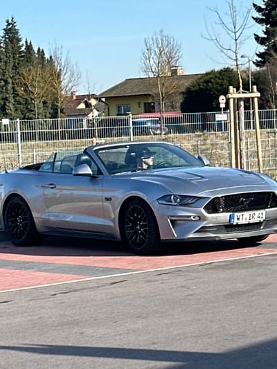 A silver Ford Mustang convertible, model year 2018 or newer, with its top retracted, is positioned on a paved area displaying red and dark gray markings. A driver, identifiable by a dark baseball cap, occupies the driver's seat. The vehicle bears a German license plate, "WT IR 41," which signifies registration within the Waldshut-Tiengen district, situating the scene in Jestetten, Germany. The Mustang features a "5.0" badge on its side panel and black multi-spoke wheels. In the background, behind a metal fence, are several residential buildings, including a light yellow house with a dark gabled roof. Bare deciduous trees and evergreen trees are visible. The sky is clear blue. A wooden utility pole, equipped with a small circular sign, stands on the right side of the frame. Shadows are cast on the foreground pavement, indicating direct sunlight.