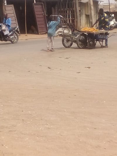 The image depicts an outdoor scene on a dusty, light brown ground surface in Tudun Murtala, Nigeria. Three individuals are present. On the far left, a man in a light blue long-sleeved shirt and blue trousers sits astride a dark-colored motorcycle, which has a white bag on its seat. In the center, a person wearing a light blue long-sleeved top and patterned light-colored trousers stands facing away from the viewer, holding the elevated handle of a two-wheeled handcart equipped with large wooden spoked wheels and carrying a light-colored sack. To the right, a man in a patterned yellow, blue, and white shirt stands behind a four-wheeled cart displaying numerous rows of orange fruits. The background features several simple structures, some with dark wooden doors or shutters and visible metal grates.