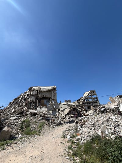 The image depicts a scene of extensive urban destruction in Jabalia, Palestinian Territory, under a clear blue sky with a sun flare visible in the upper left. The foreground features a light-colored dirt path or track, veering slightly to the right, with sparse green vegetation, including weeds and small bushes, growing along its edges and within the surrounding debris.
The middle ground and background are dominated by massive piles of rubble, remnants of what appear to be multi-story concrete buildings. On the left, a large mound of pulverized concrete, twisted rebar, and building fragments rises, with a partially intact, possibly internal, room structure visible within the debris. To the right, another significant mound of similar debris features the skeletal remains of a building, showing multiple exposed floor plates and support columns, indicative of severe structural damage and partial collapse. Exposed internal elements, such as damaged walls and fragments of personal belongings (e.g., fabric), are discernable within the shattered structures.
The entire landscape is characterized by widespread destruction, with no individuals visible within the frame. The scene is illuminated by strong sunlight, casting minimal shadows due to the overhead sun.