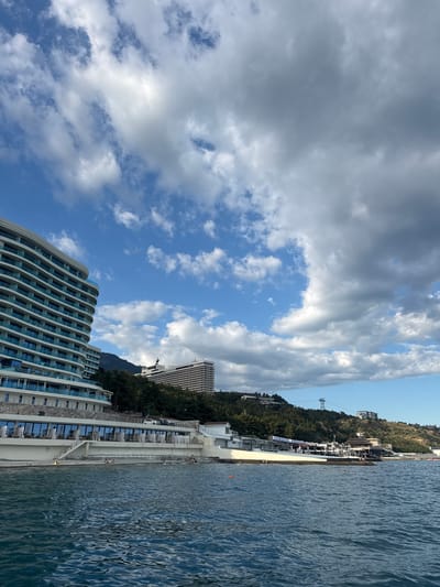 The image depicts a coastal scene in Yalta, Ukraine, with a modern hotel complex situated along the shoreline. The architecture of the hotel is striking, featuring curved lines and multiple balconies that overlook the sea.
The sea itself occupies a significant portion of the frame, with gentle waves rippling on its surface. The shoreline is visible, a narrow strip of beach leading to the water, with some people scattered along it. Behind the hotel and along the hillside, dense green foliage covers the landscape, interspersed with buildings.
In the background, a cable car can be seen ascending a hill, suggesting a connection to higher elevations and perhaps a scenic viewpoint. The sky is a mix of blue patches and dramatic, voluminous clouds, indicating partly cloudy weather. The light suggests it is daytime, likely late afternoon or early evening given the soft quality of the light.
The overall mood is serene and suggests a holiday or leisure setting. There is no visible text in the image.