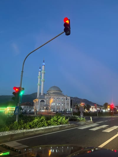 The image shows a mosque with two minarets in Köyceğiz, Turkey. The sky is a clear, deep blue, indicating it is either early morning or late evening. A tall, curving traffic light pole stands in the foreground with the top light showing red and the bottom light showing green for turning traffic. There is a crosswalk on the road to the right, with white stripes visible. A row of green bushes separates the road from the mosque grounds. The mosque is lit up with lights along its facade. Mountains are visible in the background. Street lights are visible along the road. A part of a vehicle's window is visible on the right edge of the frame.