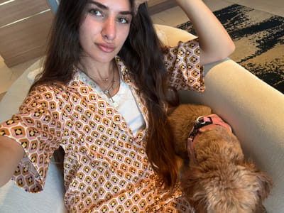 In a comfortable indoor setting, a woman with long, dark hair is seated on a beige couch, her right arm resting on her head. She wears a collared, short-sleeved shirt with a geometric pattern of pink, orange, and yellow hexagons with black and white diamond accents. Underneath, a white tank top is visible. She has a subtle smile and is looking towards the camera. Next to her, a small, fluffy, reddish-brown dog is lying down, wearing a pink harness. The dog's head is lowered, obscuring its face. The background includes a wooden wall with horizontal slats and a patterned rug with black and beige stripes. The scene appears to be in a residential living room in Boca Raton, Florida, USA.