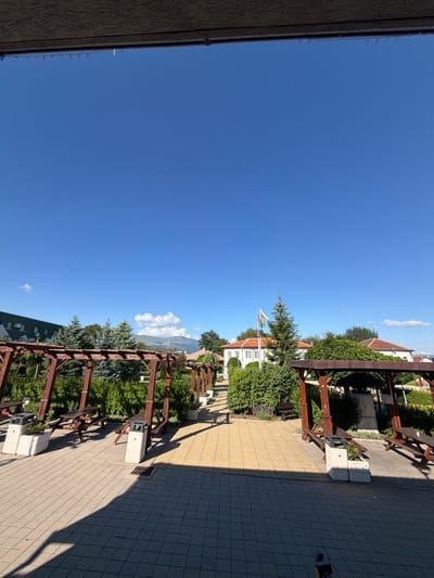 The image depicts an outdoor area with a paved walkway leading through pergolas with picnic tables and benches. The pergolas are constructed from dark wood and are covered with a lattice-like roof. Several picnic tables and benches are positioned under the pergolas, some with trash receptacles. Beyond the pergolas, there are lush green bushes and trees. In the background, white buildings with orange roofs are visible, along with a flagpole flying a flag. Further in the distance, mountains can be seen under a clear blue sky with a few scattered clouds. The overall scene suggests a park or recreational area in the municipality of Chelopech, Bulgaria.