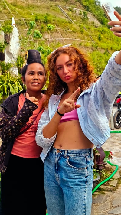 This image, captured in Tu Le, Vietnam, depicts two women posing for a selfie outdoors during daylight. On the left, a woman of apparent Southeast Asian descent, likely in her 30s or 40s, smiles directly at the camera and gives a thumbs-up gesture with her right hand. She has dark hair styled in a tall, elaborate bun with a gold-colored ornament. Her attire includes a coral-pink t-shirt, a black jacket featuring a repeating light-colored 'DG' monogram pattern on the sleeves, and black lower garments. A purple strap is visible across her chest. On the right, a woman of apparent Caucasian or Mediterranean descent, likely in her 20s or 30s, holds a light-colored smartphone with her raised left hand, taking the selfie. She has long, curly, reddish-brown hair with sunglasses perched on her head. Her expression features pursed lips, and she gestures forward with her raised right index finger. She wears a pink bandeau top, a light blue lace-textured jacket, and light blue denim jeans, exposing her midriff. A '77' tattoo is visible on her right forearm, and a black elastic band is on her right wrist. The background consists of a verdant, terraced hillside under bright sunlight, interspersed with lush green foliage. A white stone structure with surrounding plants is visible on the far left. The foreground shows a stone or paved pathway, a green hose, and parts of a motorbike on the right.