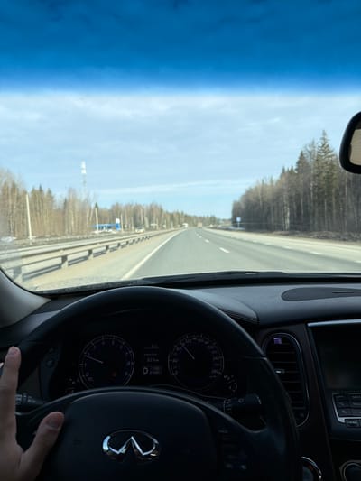 The image displays a first-person perspective from the driver's seat of an Infiniti vehicle, traversing a multi-lane highway in the vicinity of Ivanovka, Russia. A driver's left hand is positioned on the black leather steering wheel at the 9 o'clock position, featuring a silver Infiniti emblem. The instrument cluster indicates the vehicle is in 'D' (Drive), with the speedometer showing approximately 100-110 km/h and the tachometer at roughly 2000-3000 RPM. The digital odometer reads 199,026 km, with a trip meter displaying 52.1 km, and an external temperature of 5°C.
Ahead, the asphalt highway extends linearly under a partly cloudy, light blue sky. Both edges of the highway are bordered by metal guardrails and then by dense formations of leafless deciduous trees, suggesting a late winter or early spring environment. On the left side of the highway, a tall white cellular tower is prominent, alongside a small blue-roofed building and several parked vehicles, indicative of a roadside stop or service area. Multiple other vehicles are visible in the distance on the highway. The upper portion of the windshield features a dark blue tint, and a segment of the vehicle's dashboard and the rearview mirror are visible on the right.