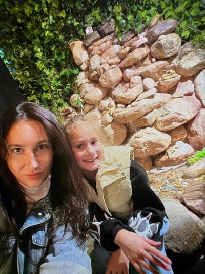 A selfie features two females in front of a rocky background with artificial greenery. The female in the foreground on the left has long, dark hair and is wearing a denim jacket over a cream turtleneck. The female to her right, who appears to be younger, has her hair in a bun and is wearing a light-colored puffer vest over a black sweatshirt with a white graphic design. She is smiling at the camera. The rocky background suggests an indoor exhibit or attraction, possibly a man-made waterfall or grotto. The lighting is artificial, creating a slightly warm tone. The setting is likely an entertainment venue within the Папярнянскі сельскі Савет, Belarus, perhaps a park or a dedicated attraction.