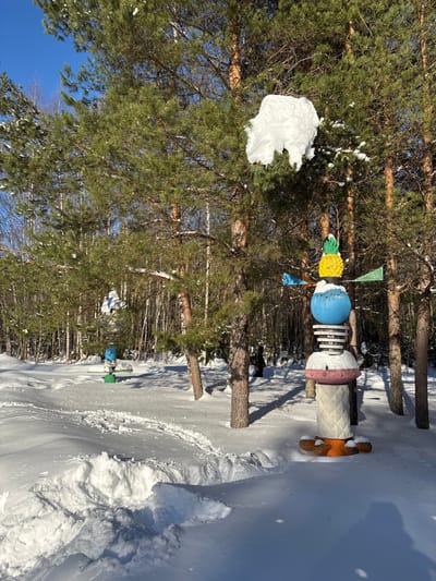 The image displays a snow-covered outdoor environment in Kol'tsovo, Russia, under a clear blue sky. A prominent multi-component sculpture stands in deep snow on the right side of the frame. This sculpture features a textured light-colored base, an orange multi-lobed section, a pink inverted dome, stacked white and dark ring elements partially covered by snow, a large blue sphere, and a yellow pineapple-shaped top with green foliage. Four small, colored fin-like vanes, including blue and green, extend radially from the blue sphere. Above and slightly behind this sculpture, a significant accumulation of snow rests on a pine tree branch. Further into the midground and to the left, a second, smaller sculpture with a blue top and green base is visible amidst the snow and trees. The ground is entirely covered in thick white snow, with a visible path of footprints leading into the scene from the lower left. Numerous pine trees with green needles and brown trunks, some holding patches of snow, populate the area, extending into a denser mixed forest in the background under bright daylight. No human figures are present.