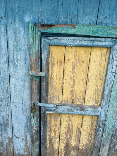 The image shows a close-up of a weathered, blue wooden structure with a small, yellow wooden door. The paint on both the structure and the door is heavily cracked and peeling, revealing the wood underneath. A rusty metal latch is attached to the blue structure, and a padlock with keys hangs from it, securing the yellow door. The wood appears aged and worn, suggesting an outdoor setting, possibly in a rural or industrial area. The overall impression is one of disuse and decay. The location is identified as Чуй-Атасево, Russia.