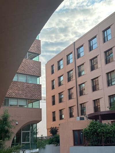The image shows the exterior of two modern buildings in Thessaloniki, Greece. The building on the left features a distinct facade with textured brickwork and large glass windows. The building on the right is a light pink, multi-story structure with numerous identical windows, each with a small black balcony railing. The sky above is partly cloudy with a soft, diffused light, suggesting it might be early morning or late afternoon. Greenery, including trees and bushes, is visible in the foreground and around the base of the buildings, indicating an urban setting with landscaping. There are no people visible in the image.