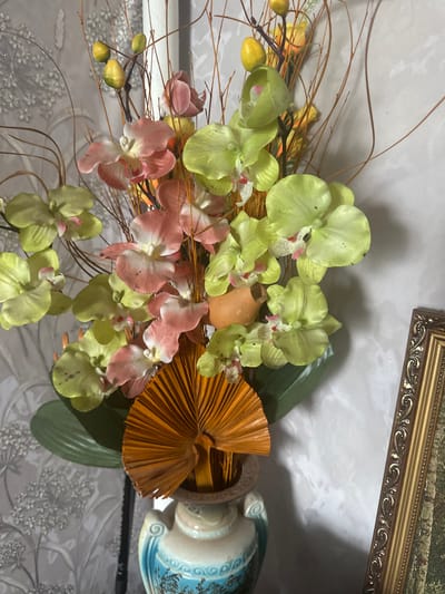 This image depicts an artificial floral arrangement in a vase. The arrangement consists of a bouquet of orchids in various shades of pink and pale green, accented with yellow bud-like elements and dry, brown twigs. A fan-shaped orange decorative element is positioned at the base of the flowers, protruding from the vase. The vase itself is ceramic, with a light blue and white patterned design. The background features a textured wallpaper with a light gray floral pattern. A portion of a framed picture with an ornate golden frame is visible on the right side of the image. The scene appears to be indoors, likely in a residential setting. There are no people or discernible actions taking place. The location of Yazykova Pyatina, Russia, is not visually represented or implied by the image content.
