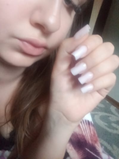 The image is a close-up, low-angle, portrait-style photograph of a young person's face and hand. The person's face is partially visible, with their mouth and nose in focus. Their lips are closed and appear to be slightly parted. They have light-colored skin and long, dark brown hair. Their fingernails are painted a solid, light color, possibly white or a very pale pink, and appear to be artificial or significantly elongated. The hand is positioned in front of the face, with fingers slightly curled. The background is blurred and appears to be an indoor setting, possibly a room with a patterned rug or blanket visible in the lower right corner. There is no visible indication of the location within Elizaveta, Moldova.