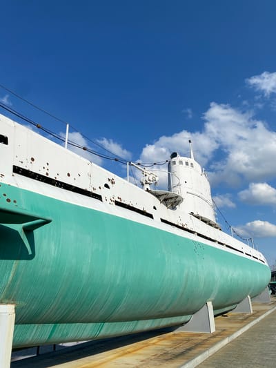 The image shows a decommissioned submarine, painted in white and teal, resting on a display stand in the city of Verkhnyaya Pyshma, Russia. The submarine is positioned outdoors under a clear blue sky with scattered white clouds. The hull of the submarine exhibits signs of wear, with rust spots visible on the white upper section. A periscope and other external structures are mounted on the conning tower. The submarine appears to be part of a museum or memorial, as it is supported by sturdy grey stands and situated on a paved area, possibly an exhibition ground. The background is dominated by the expansive sky, with hints of greenery and other structures visible in the far distance, suggesting an outdoor, possibly urban or industrial, setting.